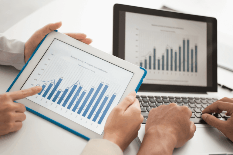 Duas pessoas analisando gráficos de desempenho empresarial em dispositivos digitais, com uma pessoa apontando para um gráfico de barras em um tablet e outro gráfico similar sendo exibido em um laptop, é a imagem que ilustra artigo sobre Dynamics 365.