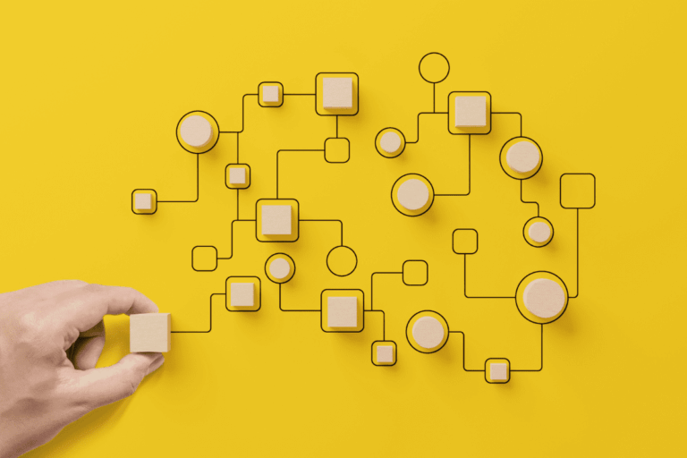 ilustrando artigo de introdução ao SharePoint, temos a representação visual de um diagrama de conexões em fundo amarelo, com linhas pretas interligando blocos e círculos de madeira, simbolizando organização, fluxo de trabalho e conectividade. Uma mão posiciona um dos blocos, indicando interação ou montagem.