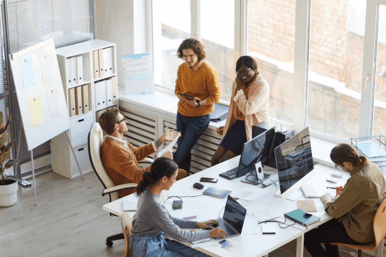 foto que ilustra artigo sobre dicas de produtividade com Microsoft Loop: grupo de cinco pessoas trabalhando em um escritório iluminado pela luz natural. Três pessoas estão em pé, próximas a uma janela, interagindo e sorrindo, enquanto outras duas estão sentadas em volta de uma mesa. Uma mulher com laptop parece concentrada, e outra pessoa faz anotações em um caderno. O ambiente é moderno, com mesas brancas, monitores de computador e uma estante com pastas organizadas ao fundo. A atmosfera parece descontraída e colaborativa.