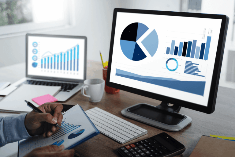 Foto ilustrando artigo sobre Power BI. Pessoa analisando gráficos em uma tela de computador, com outro laptop ao lado exibindo dados em formato de gráfico de barras e de linha. A imagem também mostra a pessoa segurando uma prancheta com anotações de gráficos, indicando um ambiente de trabalho focado em análise de dados.