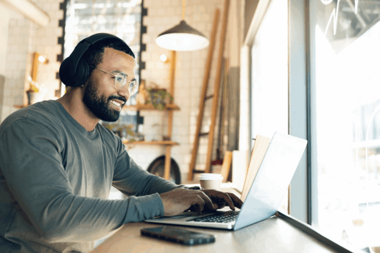 homem sorridente trabalhando em um laptop em um ambiente de café, usando fones de ouvido e óculos, com uma xícara de café ao lado, é foto que ilustra artigo sobre cibersegurança no trabalho remoto.