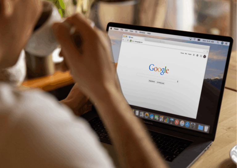Representando as novidades do Google Authenticator, a fotografia mostra o foco em um notebook, com uma pessoa sentada a frente dele e bebendo em uma xícara de café, a tela exibe a página principal do Google, em um ambiente de home office.