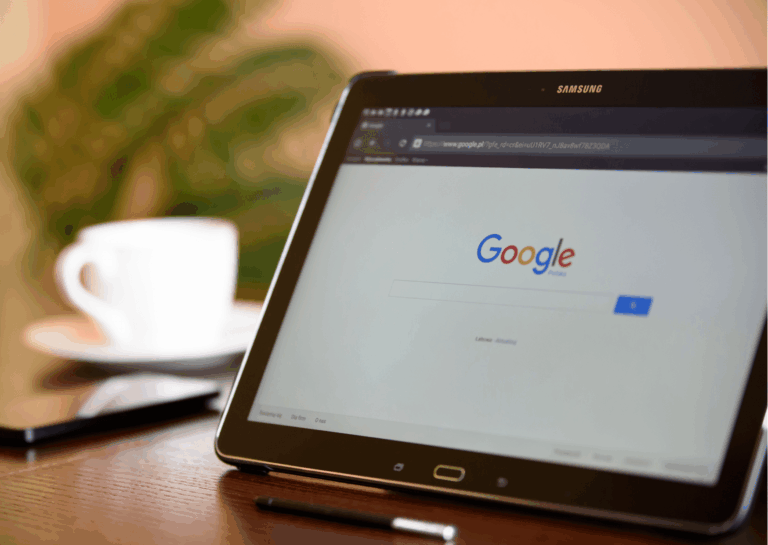 Um tablet com caneta está sobre uma mesa de madeira, deitado verticalmente e exibe a tela de pesquisa do Google, ao fundo é possível ver, em desfoque, uma xícara branca com pires e um celular; exemplificando o Google Passkey.