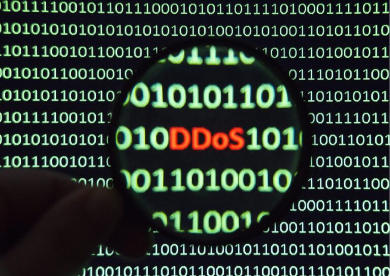 Representando o que é um ataque DDos, a imagem mostra sobre um fundo preto, linhas de códigos binários em cor verde, e, sobre ela, aparece a parte de uma mão segurando uma lupa, que dá ênfase ao escrito DDoS, na cor vermelha, no meio de uma das linhas de numerais zero e um.