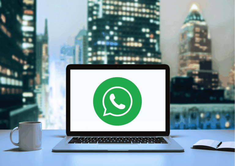Em um ambiente de trabalho, com foto de cidade ao fundo, está um notebook, uma caneca e um caderninho aberto em cima de uma mesa e, na tela do notebook, está uma logo do aplicativo WhatsApp para Windows.