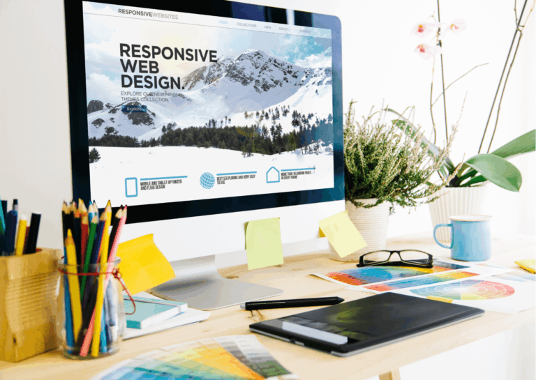 Sobre uma mesa de escritório, colocada rente à uma parede branca, está um desktop branco com borda da tela preta e, na tela, aparece a imagem de uma montanha, aparentemente um site de teste, com título em destaque escrito "responsive web design"; na frente do desktop e ao redor, estão espalhados materiais de escritório, como porta-lápis, prancheta, papéis, óculos, caneca, post-its na tela, cadernetas, caneta, etc., e, ao fundo e canto direito, algumas plantas e orquídeas; exemplificam o que é um plugin.
