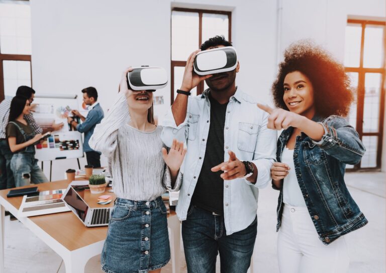 Representando os benefícios da realidade virtual para empresas, a fotografia mostra um ambiente de trabalho com mesa ao fundo e uma equipe trabalhando em um moodboard, e três pessoas em destaque à frente, duas delas utilizando óculos de realidade virtual e uma delas sem óculos, explicando seu uso.